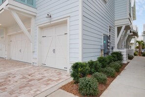 A residential building with two white garage doors, a brick driveway, and a landscaped garden with shrubs and a palm tree. - Beach | Bikes | Great Location | Amazing Pools (Santa Rosa Beach)
