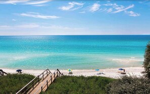 A beach with a wooden walkway leading to the shore, clear blue water, and people relaxing under umbrellas. - Beautiful Condo | Private Beach Access | Two Pools (Santa Rosa Beach)