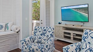 Una sala de estar con un televisor grande que muestra una vista costera, un sillón estampado, un banco blanco con cojines y un aparador de madera.