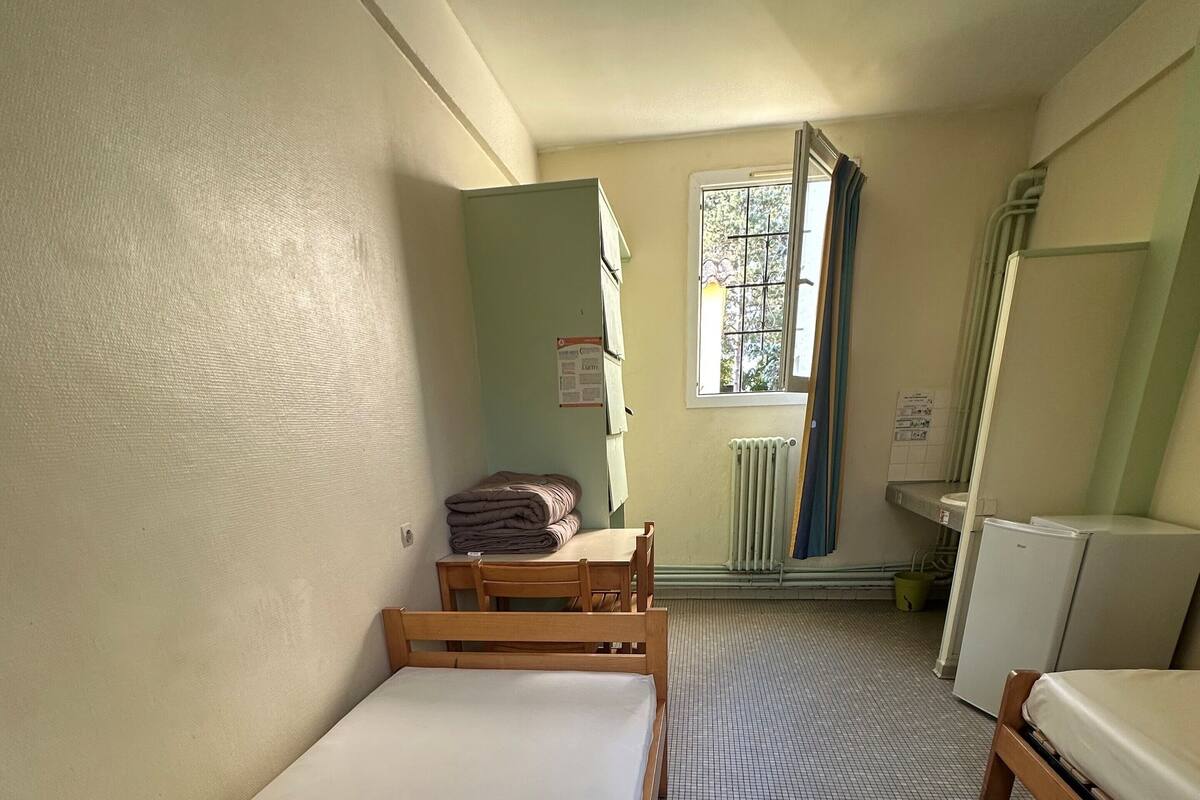 Una habitación pequeña y funcional con una cama individual, una mesa de madera y una ventana con cortinas.