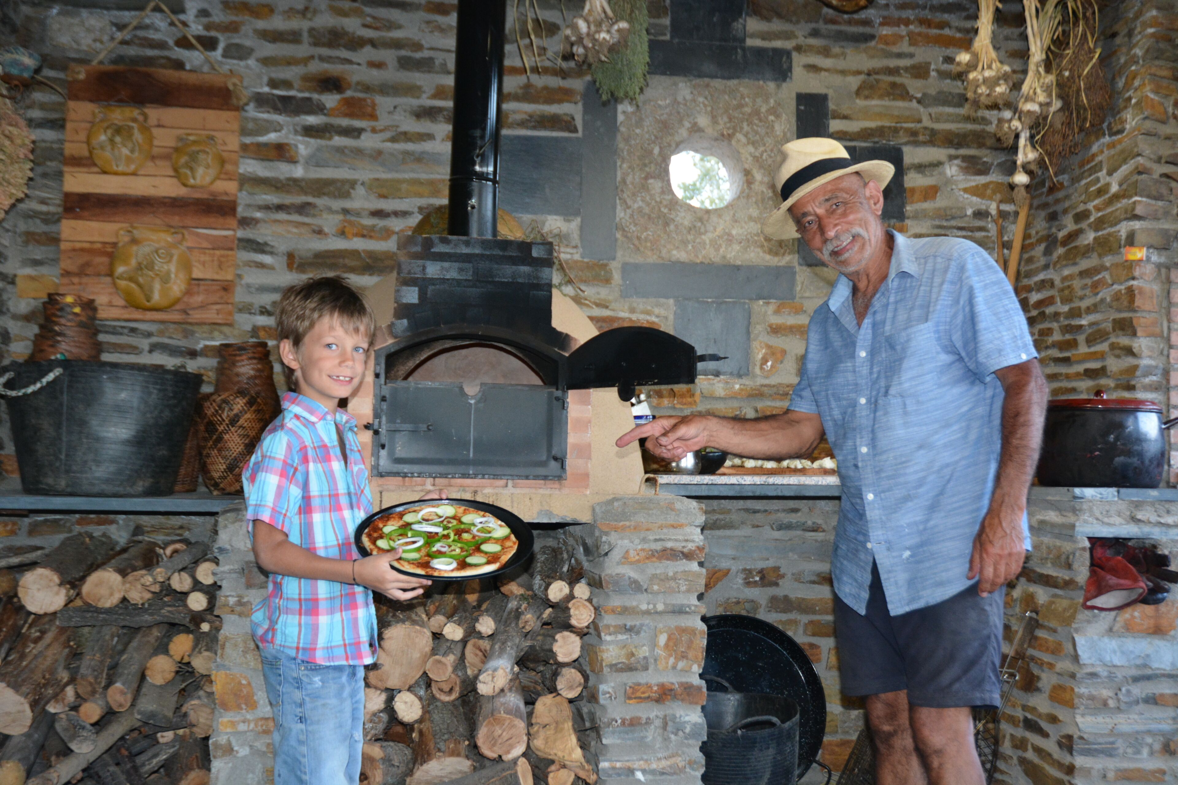 A stone fireplace with a pizza oven, a young boy holding a pizza, and an older man wearing a hat.