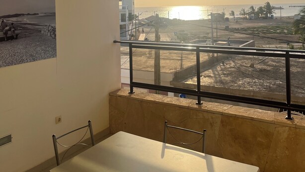Un balcon avec une table et des chaises, donnant sur un chantier de construction et offrant une vue sur la mer et les bâtiments au loin.