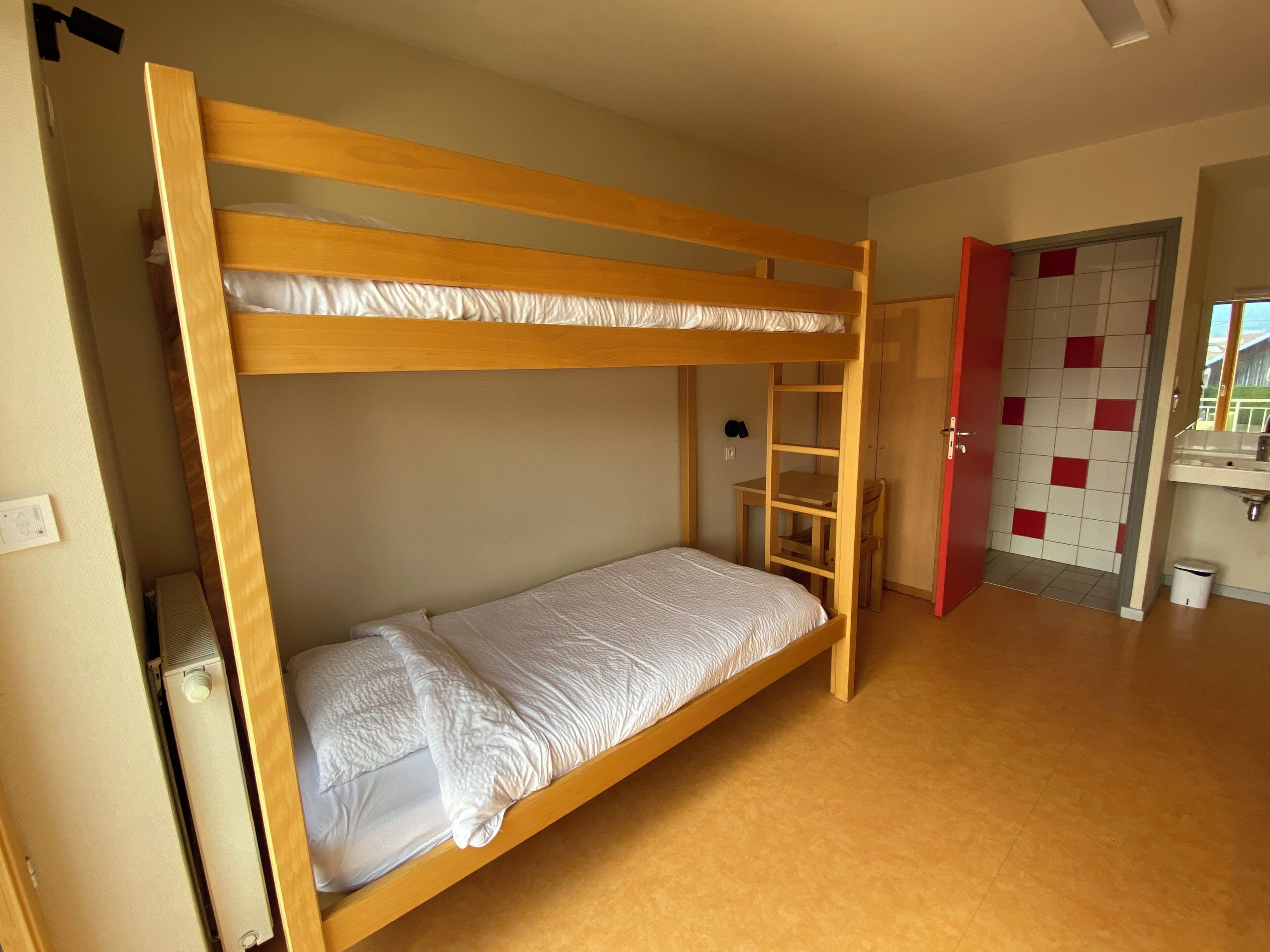 Une chambre avec un lit superposé, une échelle en bois, une petite table et une porte donnant sur une salle de bain carrelée de rouge et de blanc.