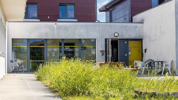 A modern building with large windows and a yellow door, surrounded by greenery and outdoor seating.