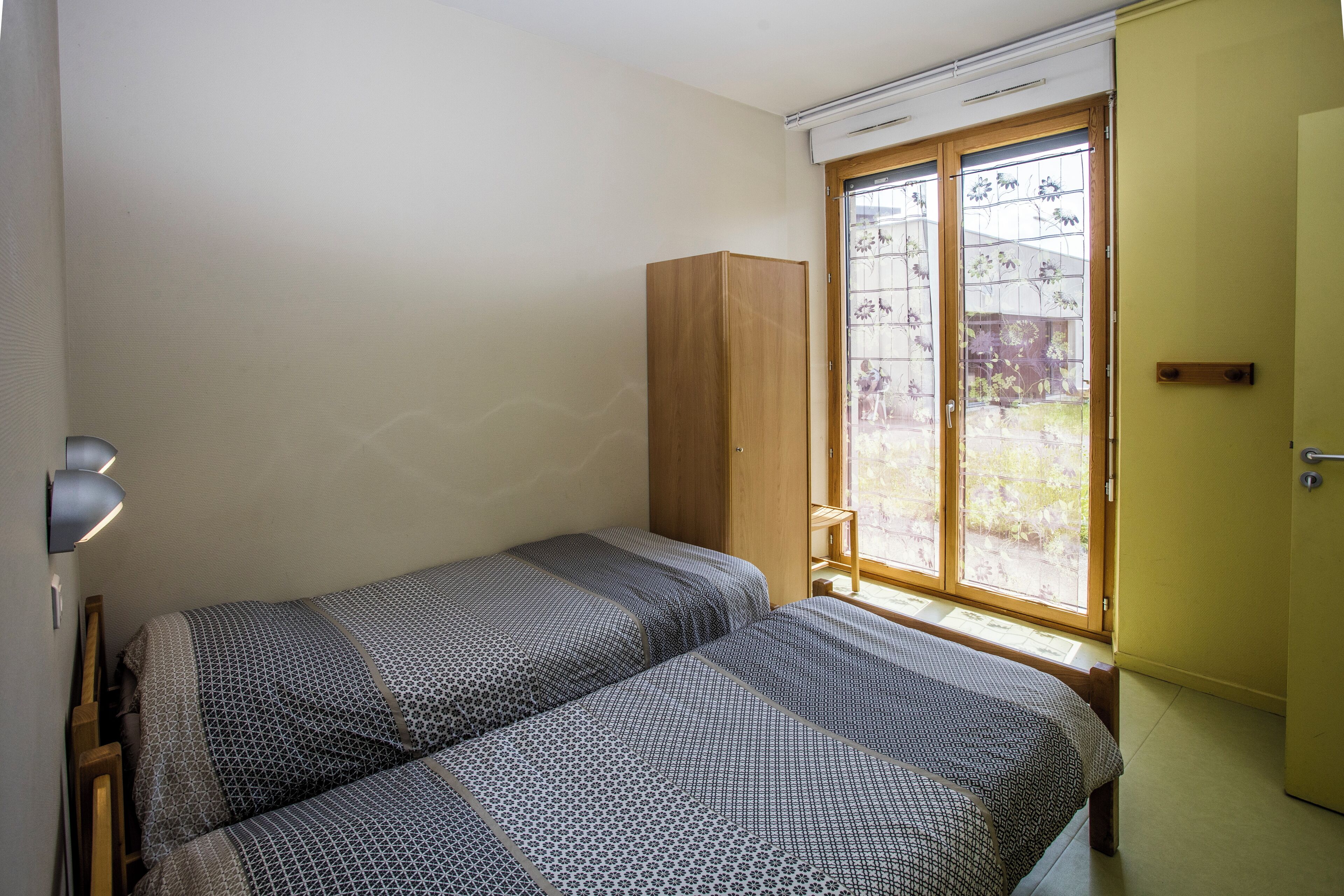 Un petit logement composé d’une seule pièce, comprenant deux lits simples, une armoire en bois, une fenêtre donnant sur un espace verdoyant et une porte.