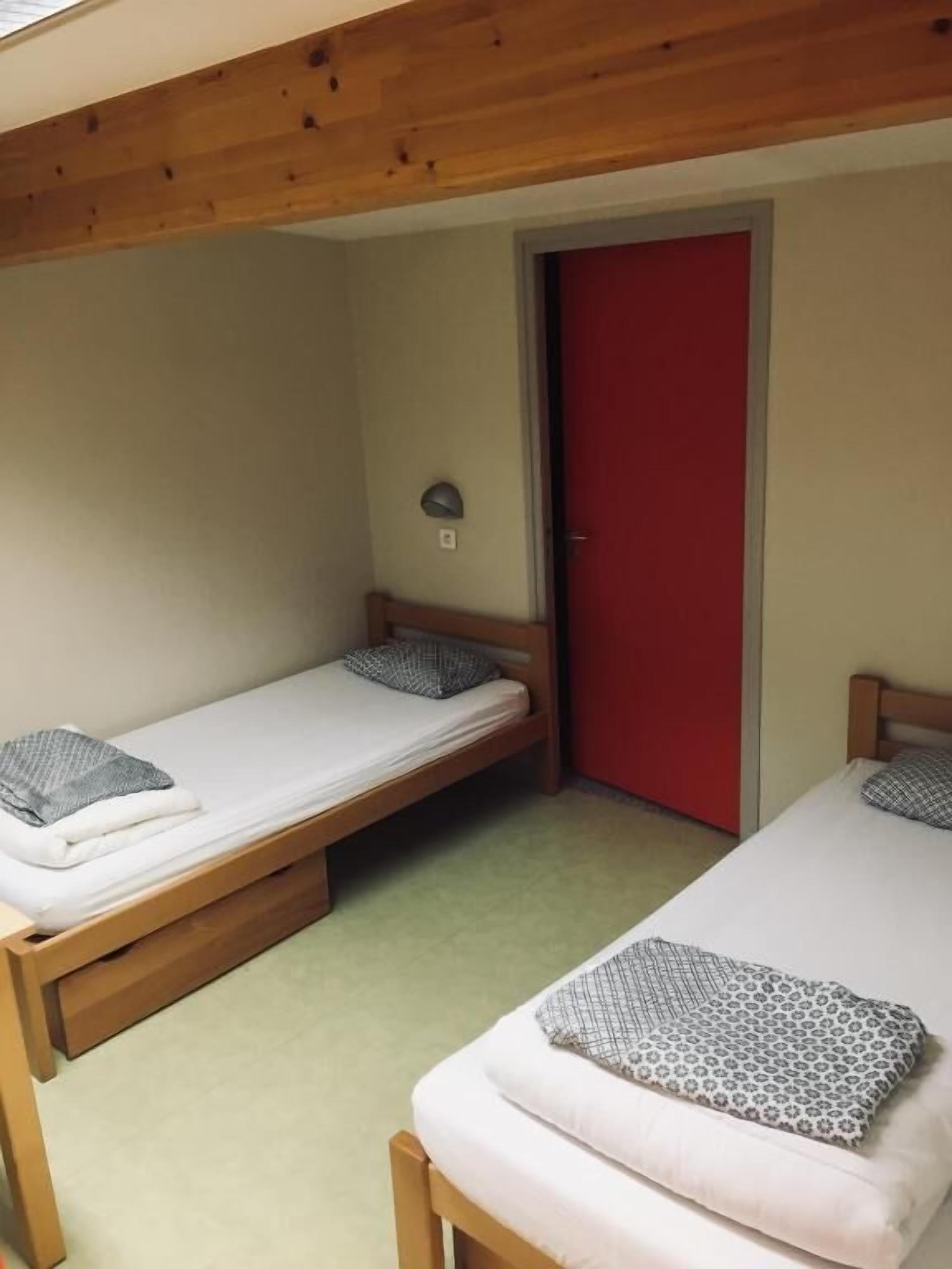 Una habitación pequeña y funcional con dos literas de madera, una puerta roja y una lámpara pequeña en la pared.