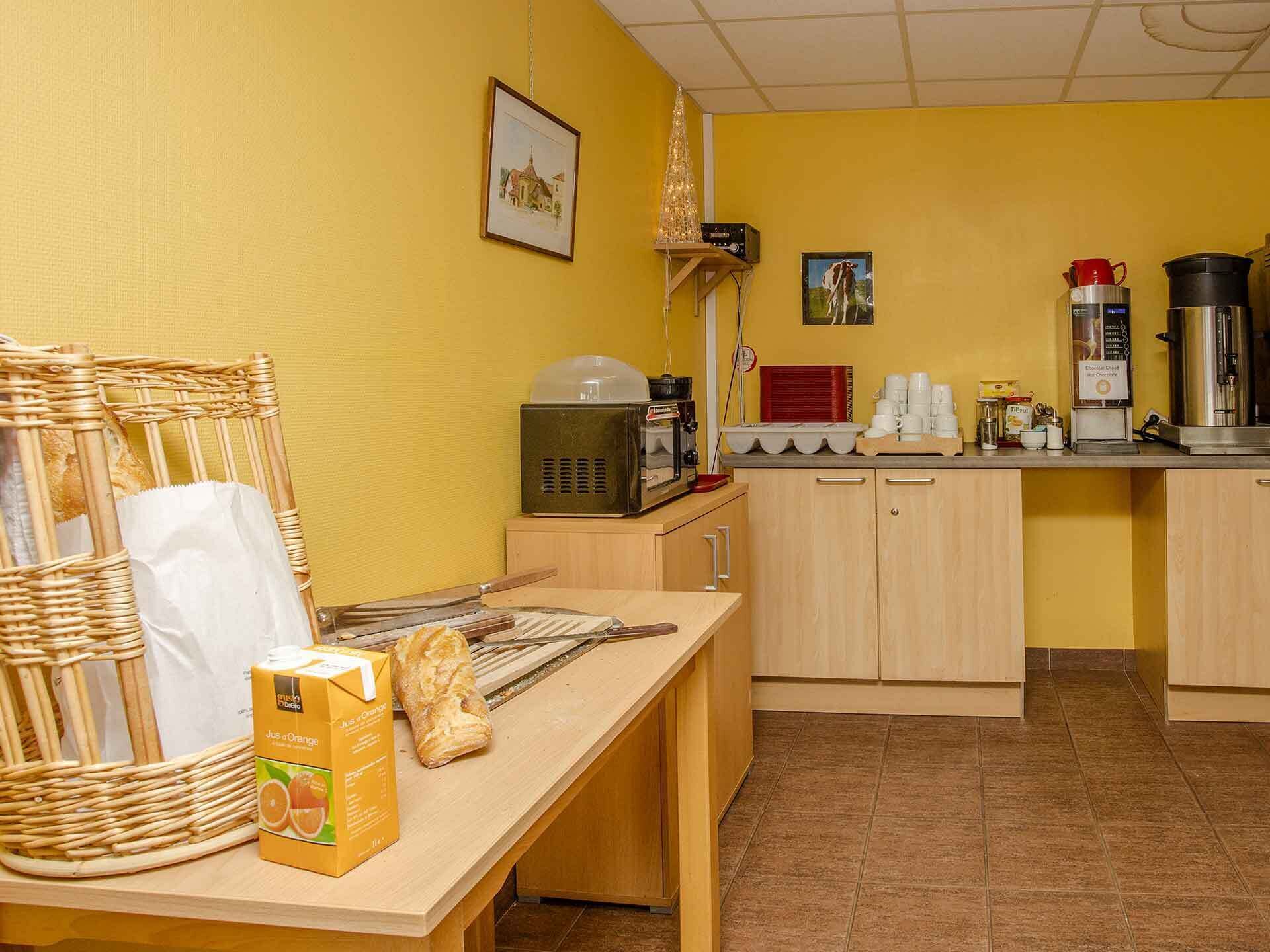 A breakfast area with a wicker chair, a table with a carton of orange juice, a loaf of bread, and a coffee maker.