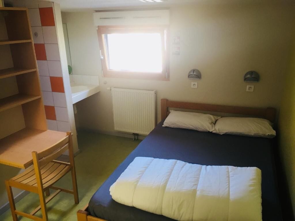 Un dormitorio con cama, una silla, estantería, ventana y radiador.