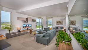 Una sala de estar moderna con un sofá seccional gris grande, un televisor de pantalla plana y vistas a un paisaje verde a través de amplios ventanales.