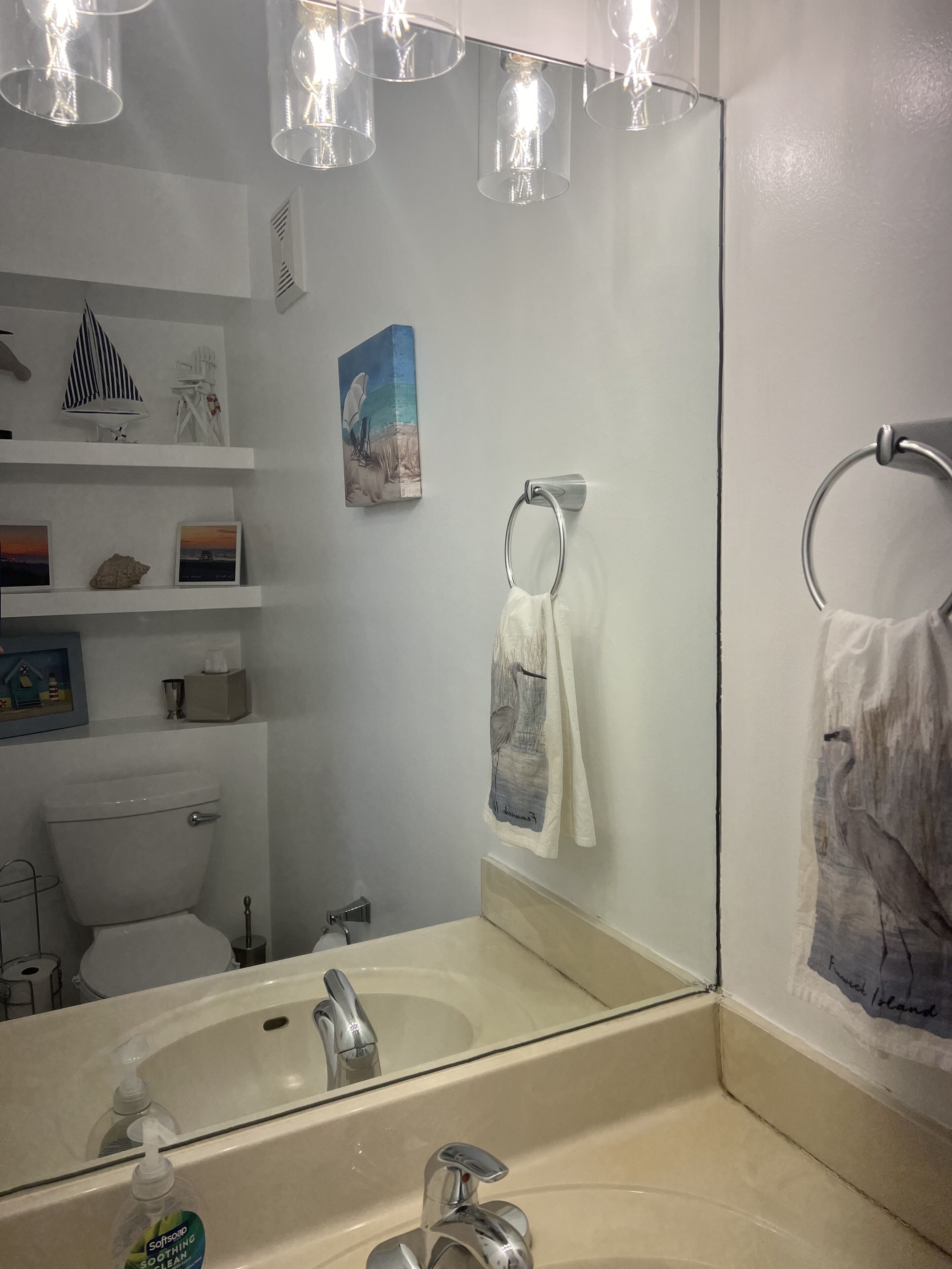 A bathroom with a toilet, sink, and towel rack. There are shelves with decorative items and a painting on the wall.