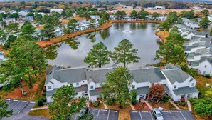 An aerial view of a residential area with houses surrounding a central lake, surrounded by trees and open fields. - Mallard Lakes Waterfront Beach Condo in Fenwick Island with Two Pools (Selbyville)