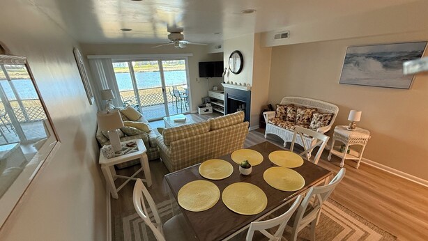 A dining area with a wooden table and white chairs, a sofa, and a television in the background. - Mallard Lakes Waterfront Beach Condo in Fenwick Island with Two Pools (Selbyville)