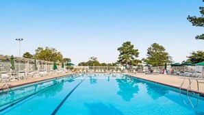 A clear blue swimming pool with a brick border, surrounded by white lounge chairs and umbrellas, and trees in the background. - Mallard Lakes Waterfront Beach Condo in Fenwick Island with Two Pools (Selbyville)