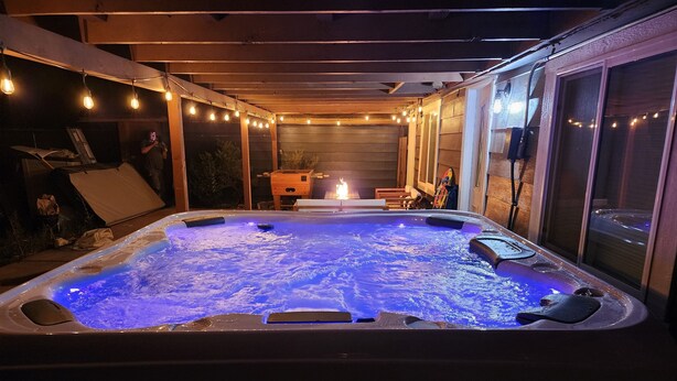 Un jacuzzi iluminado con luz azul, rodeado por una terraza de madera y luces colgantes.