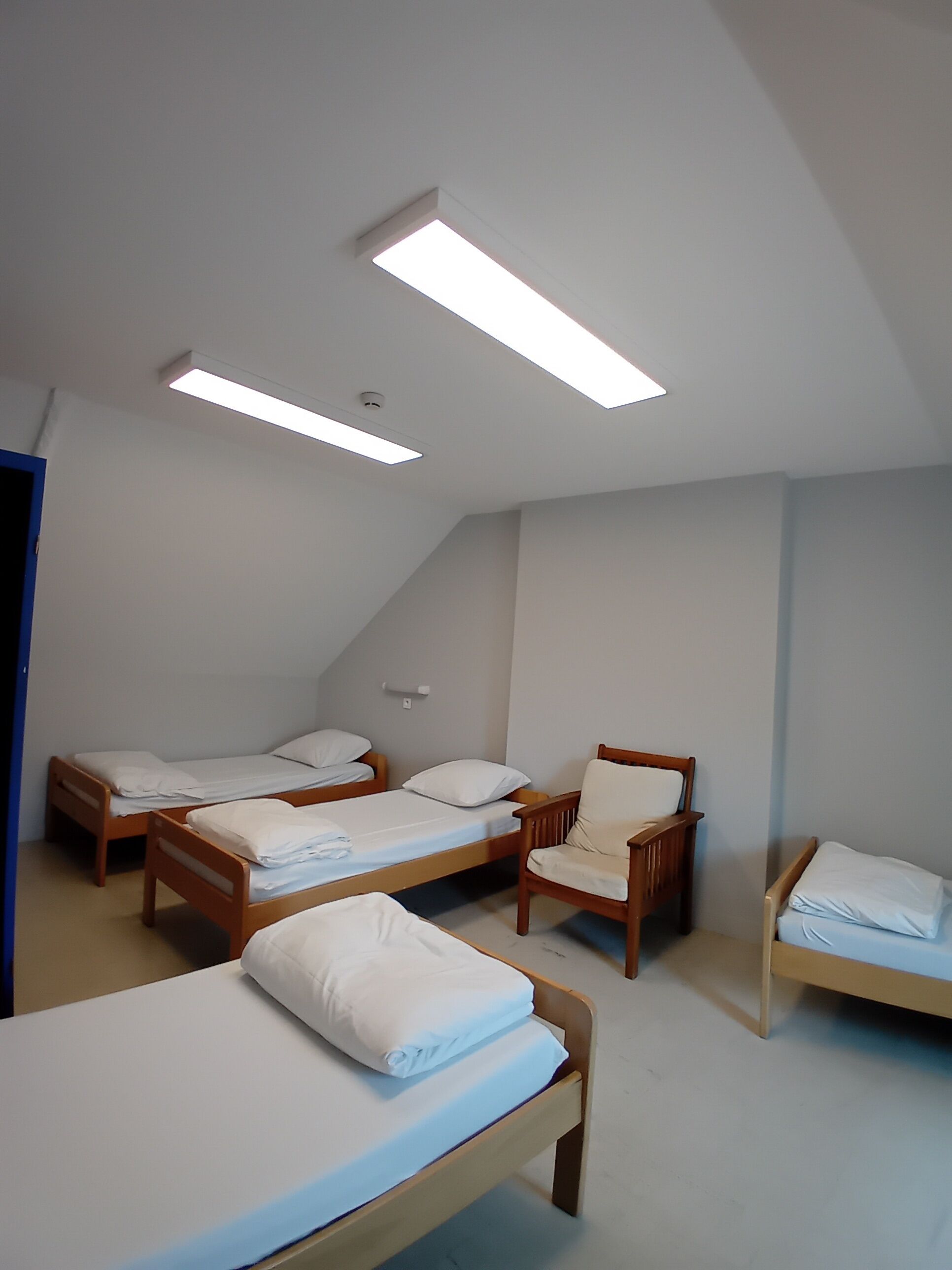 A dormitory room with four single beds, a wooden chair, and a ceiling light fixture.