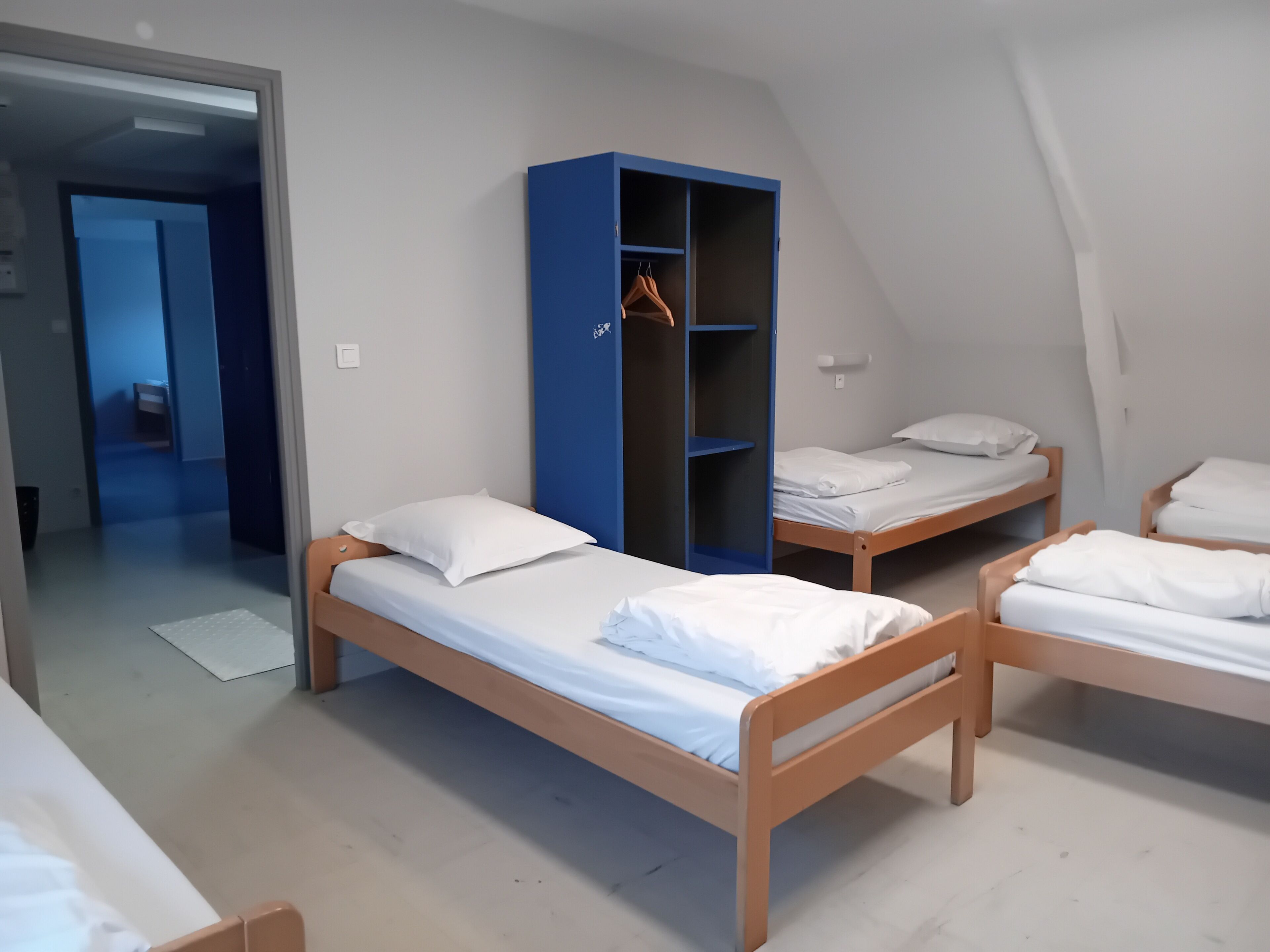 A dormitory room with three wooden bunk beds, a blue wardrobe, and a white door leading to another room.