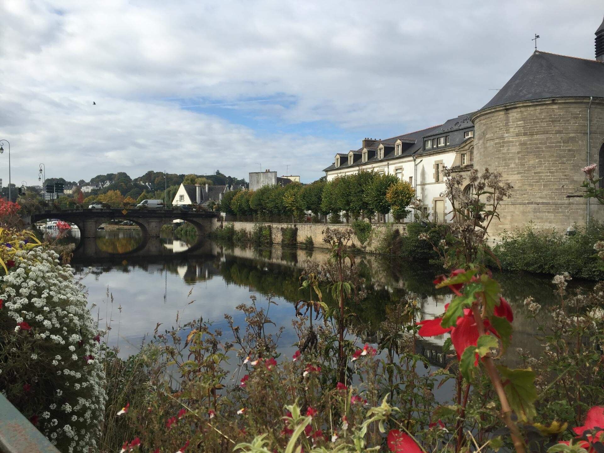 A historic building with a round tower, a bridge, and a calm river reflecting the structure.