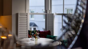Ein Zimmer mit einem runden Tisch, einer Weinflasche und Gläsern darauf. Auf der Couch liegen bunte Kissen und es gibt ein Fenster mit Jalousien.