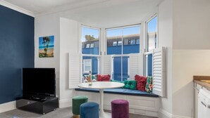 Ein Zimmer mit Erkerfenster, Flachbildfernseher, rundem Tisch und bunten Poufs.