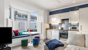 Eine moderne Wohnung mit Küche, Essbereich und Wohnzimmer. Sie verfügt über ein großes Fenster, einen Flachbildfernseher und ein Sofa mit Überwurfdecke.