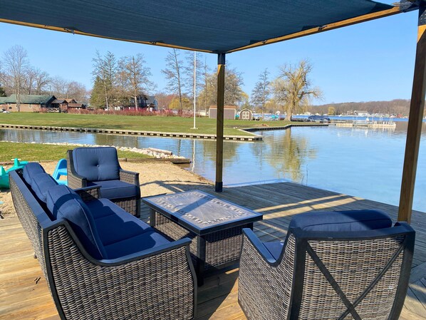 An outdoor deck with wicker furniture, a table, and a view of a lake and buildings in the background.