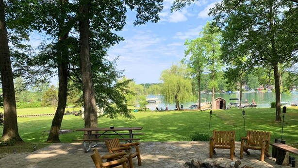 An outdoor seating area with wooden chairs and tables, a fire pit, and a view of a lake with boats. - Charming Lakefront Cottage (Vandalia)