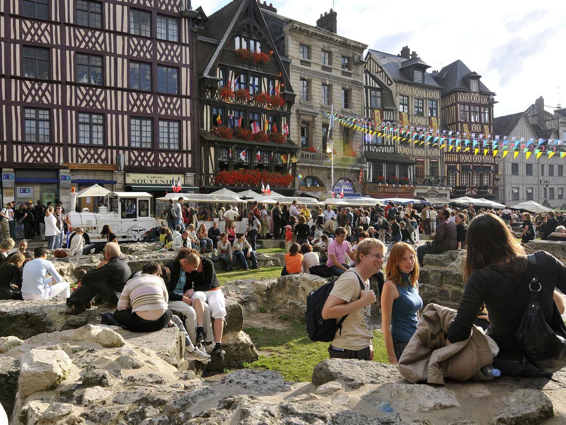 Ein belebter Markt im Freien mit steinernen Sitzgelegenheiten und traditioneller europäischer Architektur im Hintergrund.