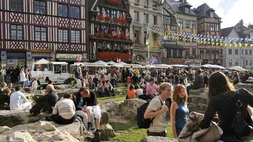 Un marché animé en plein air, avec des bancs en pierre et, en arrière-plan, des bâtiments d’architecture européenne traditionnelle.
