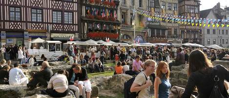 Ein belebter Markt im Freien mit steinernen Sitzgelegenheiten und traditioneller europäischer Architektur im Hintergrund.