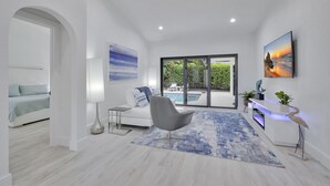A modern living room with a white sofa, a gray armchair, a blue rug, a flat-screen TV, and a pool view.