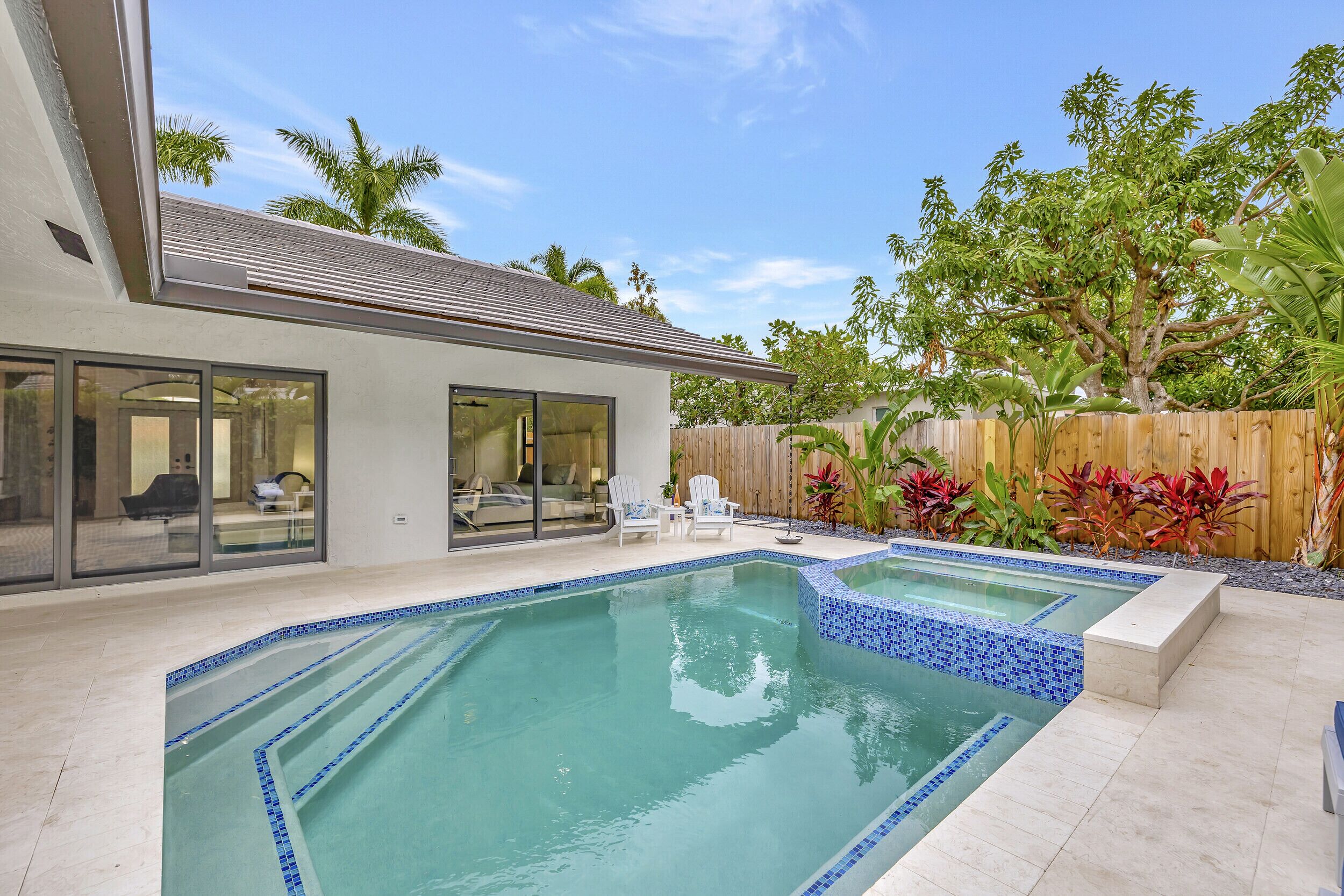 A swimming pool with a tiled edge, a wooden fence, and tropical plants.