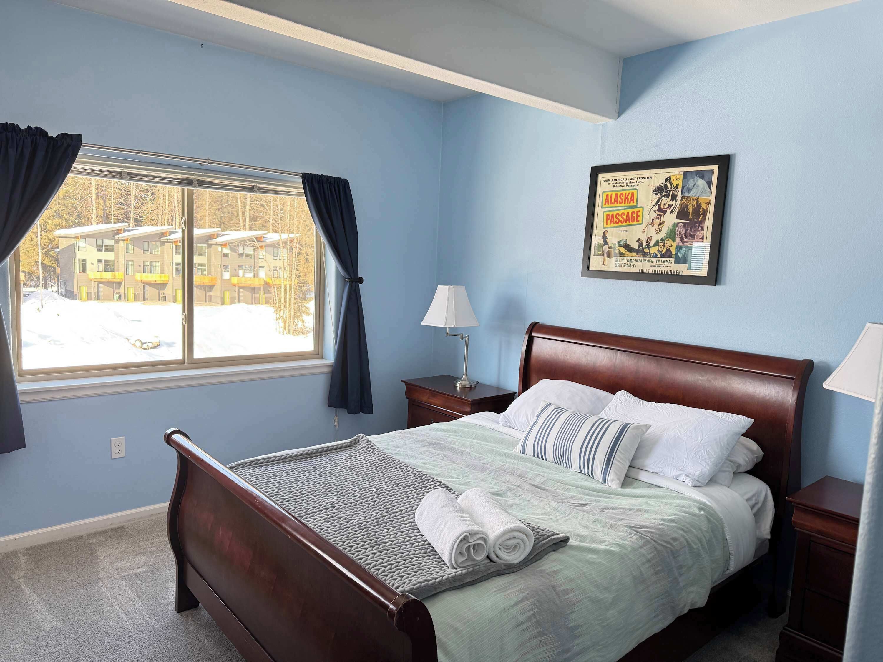 A bedroom with a bed, a window with curtains, a nightstand, a lamp, and a framed picture on the wall.