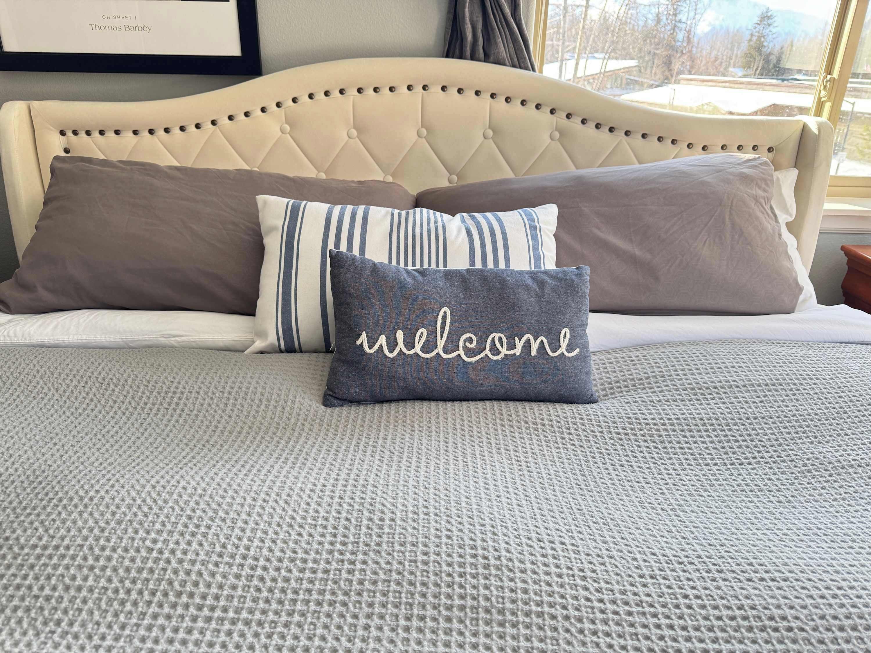 Una cama con cabecera capitonada, un edredón gris con textura y dos almohadas, una de ellas con el bordado 'welcome'.