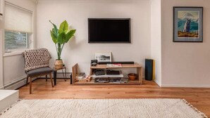 A living room with a flat-screen TV mounted on the wall, a wooden entertainment unit with books and a plant, a chair with a blanket, and a framed picture on the wall. - 2 Bedroom Modern Condo in Heart of Downtown (Anchorage)