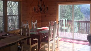 A wooden cabin with a dining table, chairs, and a chandelier. There are large windows and a balcony with a view of trees.