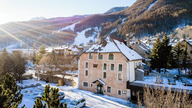 A building with a snow-covered roof, surrounded by trees and a mountainous backdrop.