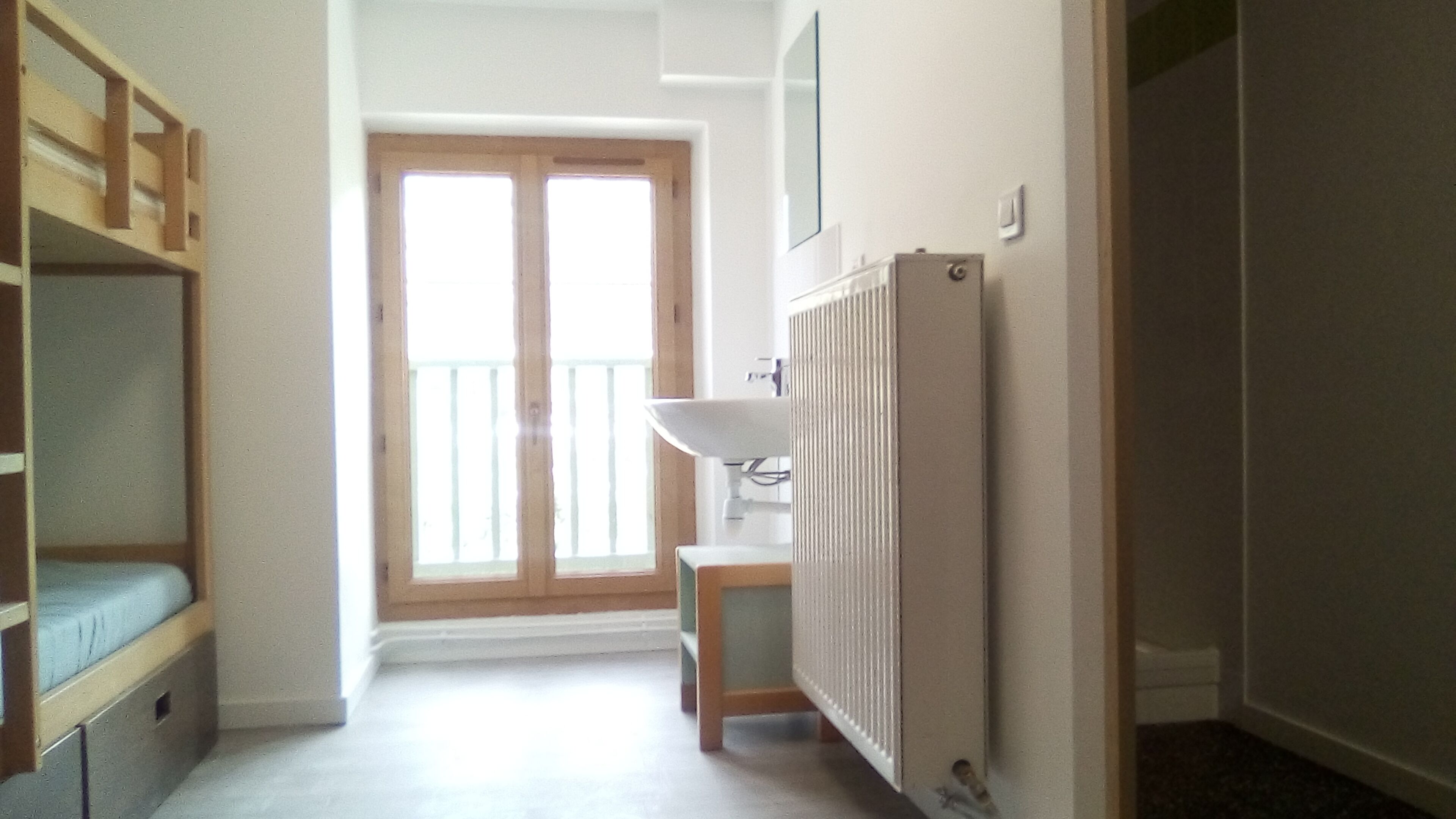 A room with a wooden bunk bed, a small table, a radiator, and a window with a view of a balcony.