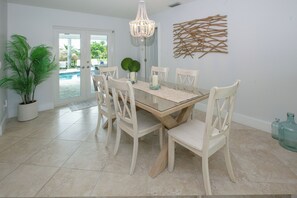Comedor con mesa de vidrio, sillas blancas, una lámpara de araña y vista a una piscina a través de puertas de cristal.