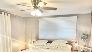 Un dormitorio con cama de madera, ventilador de techo, mesita de noche con lámpara y ventana con persianas.