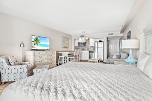 Une chambre d’hôtel avec un grand lit, un bureau avec une chaise, un coin cuisine et un téléviseur diffusant des images d’une plage.