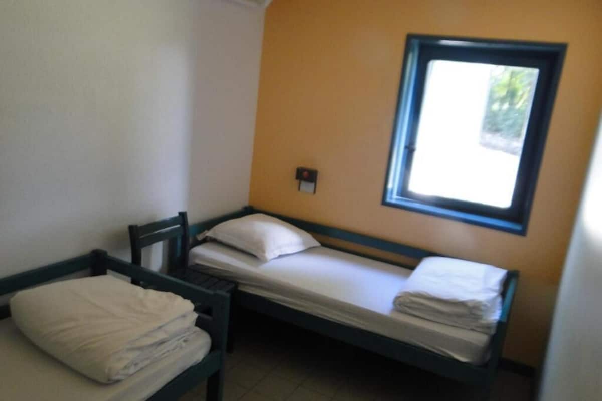 A small, single-bed room with a window, a bedside table, and a chair.