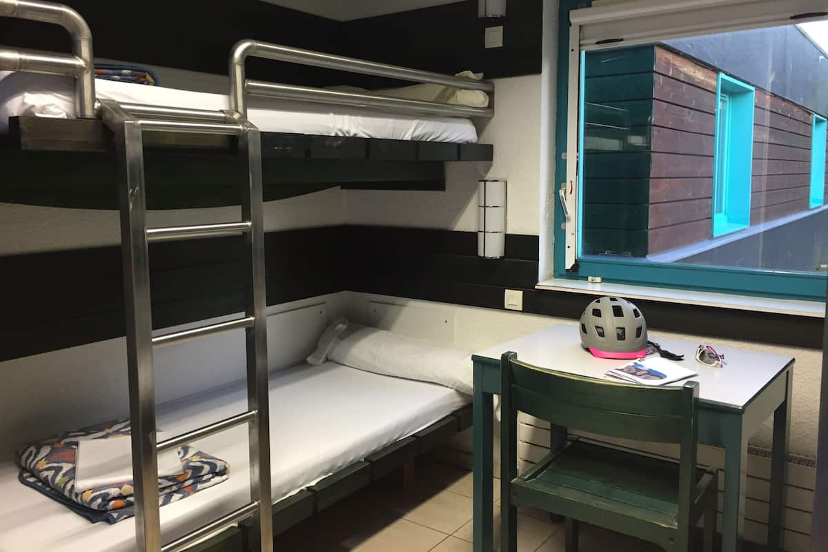 A bunk bed room with a desk and chair, a window with a view of buildings, and a helmet on the desk.