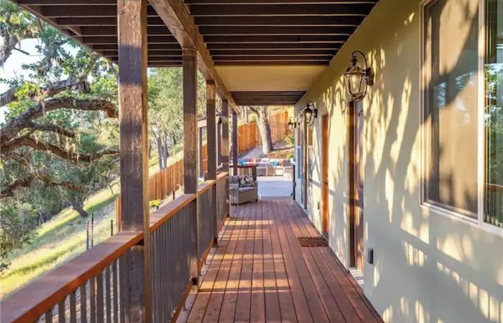 A covered wooden deck with a railing, a bench, and a view of a backyard with trees and a fence.
