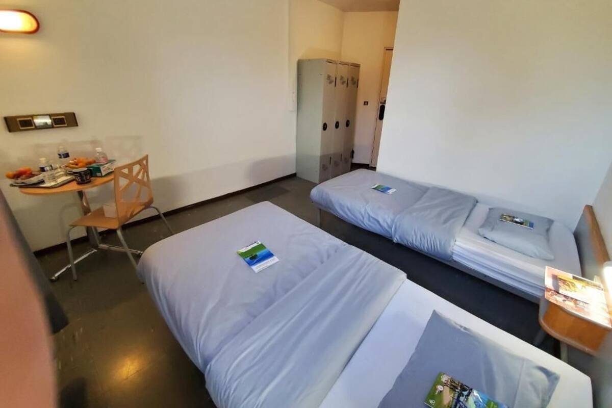 A hotel room with two single beds, a desk with a chair, a wardrobe, and a small table with a lamp.