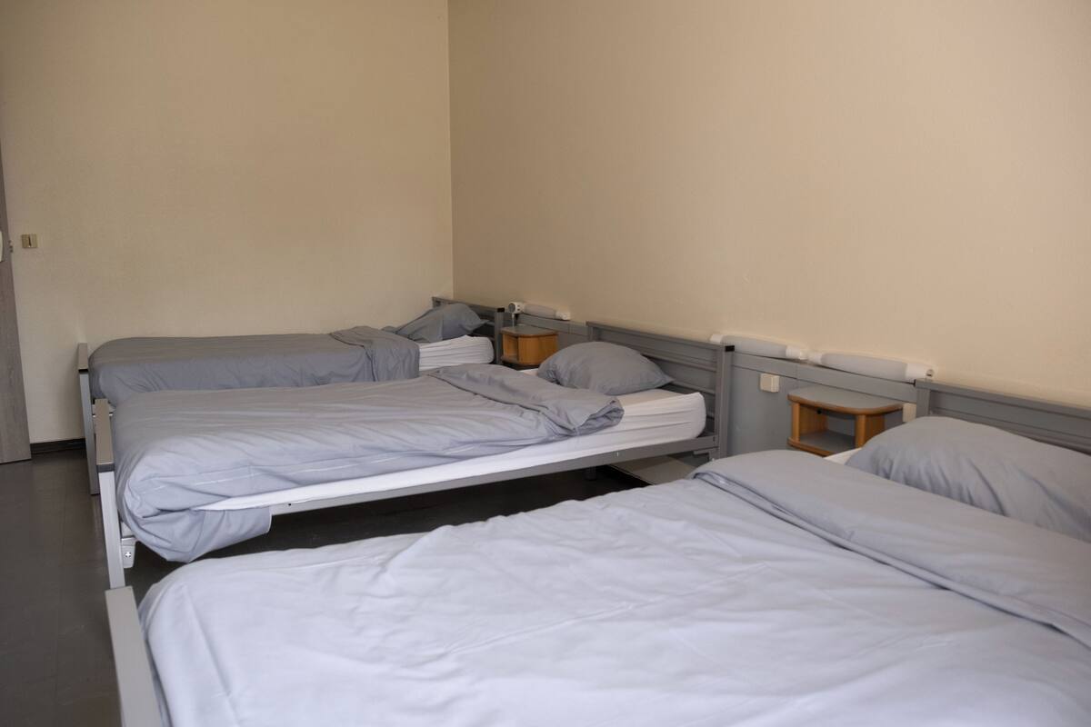 A room with three single beds, each with a grey mattress and pillow. There is a small wooden nightstand with a lamp on one side.