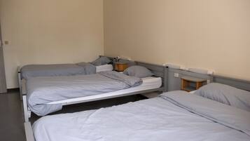 Een kamer met drie eenpersoonsbedden, elk met een grijze matras en kussen. Aan één kant staat een klein houten nachtkastje met een lamp.
