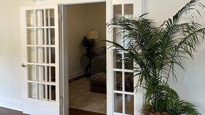 Un umbral abierto que da a un dormitorio con una cama, una mesita de noche y una lámpara. Una planta en maceta está en primer plano.