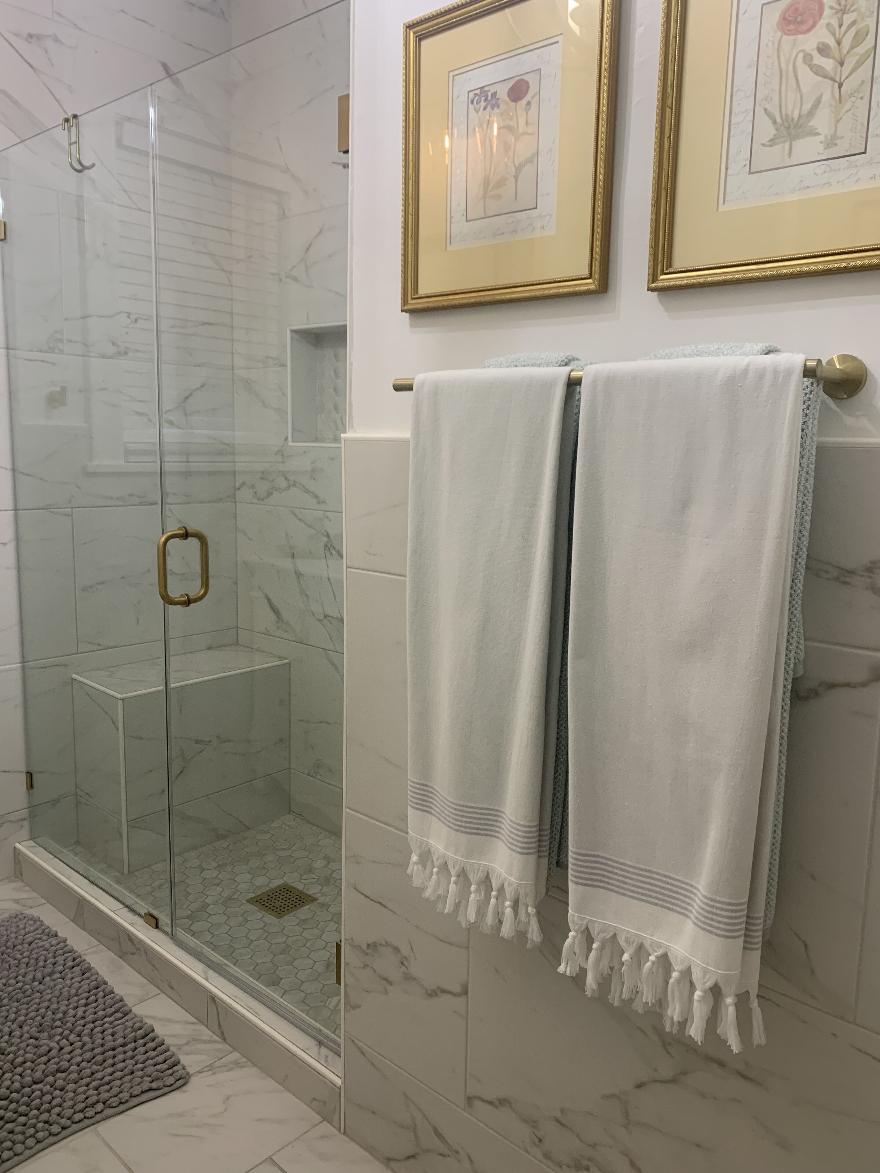 Un baño con cabina de ducha de vidrio, azulejos de mármol y dos láminas botánicas enmarcadas en la pared.