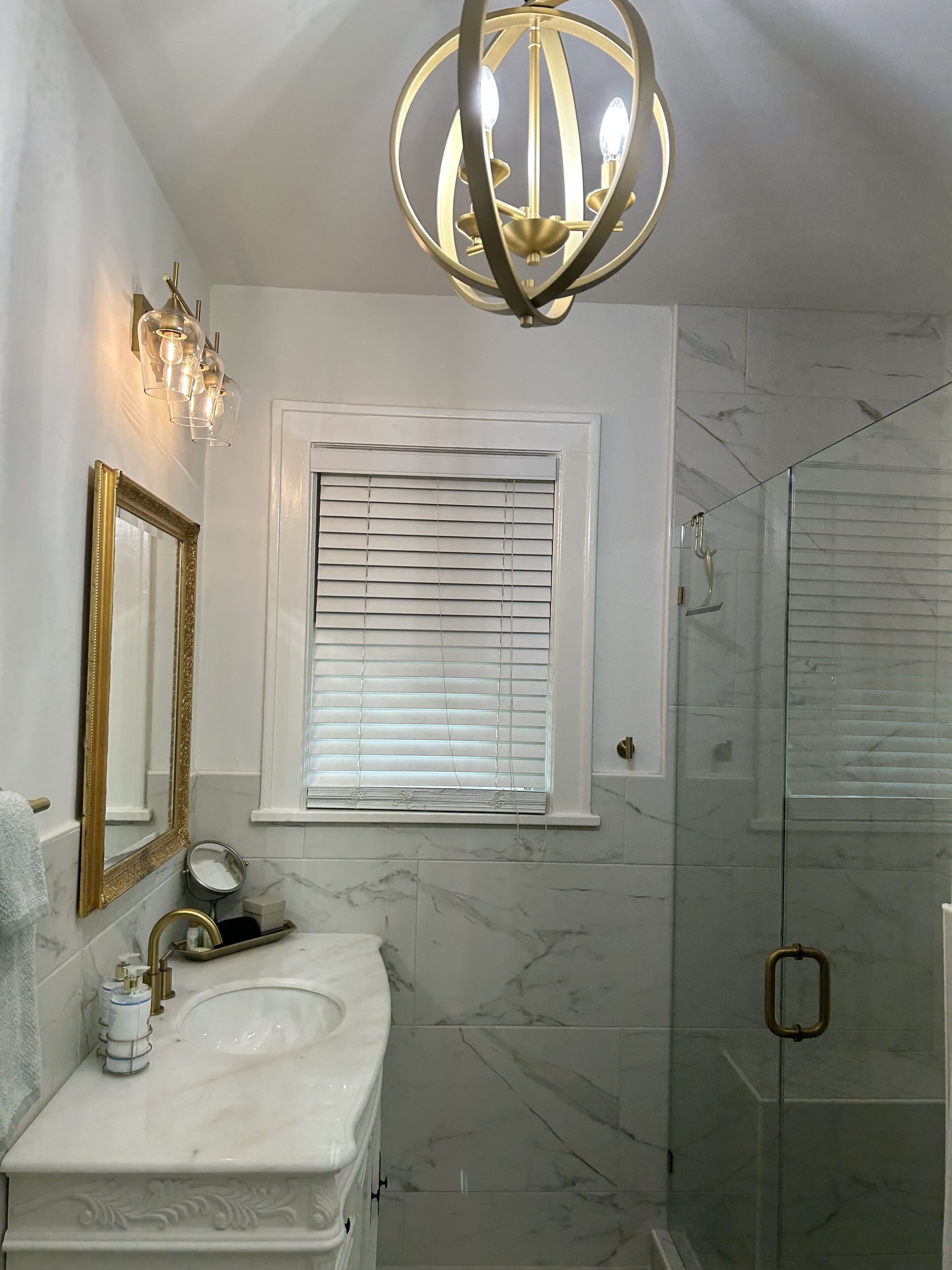 Un baño con lavamanos de mármol, espejo con marco dorado, candelabro y ducha con mampara de vidrio.