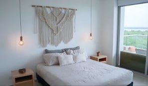 Quarto com cama, mesas de cabeceira, um painel de macramé na parede e uma janela grande com vista.
